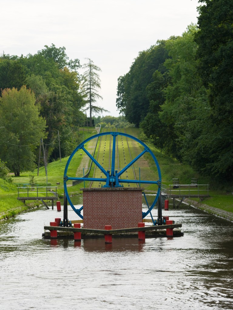Widok na koło wodne umieszczone w kanale, otoczone zielonymi drzewami i trawnikiem.