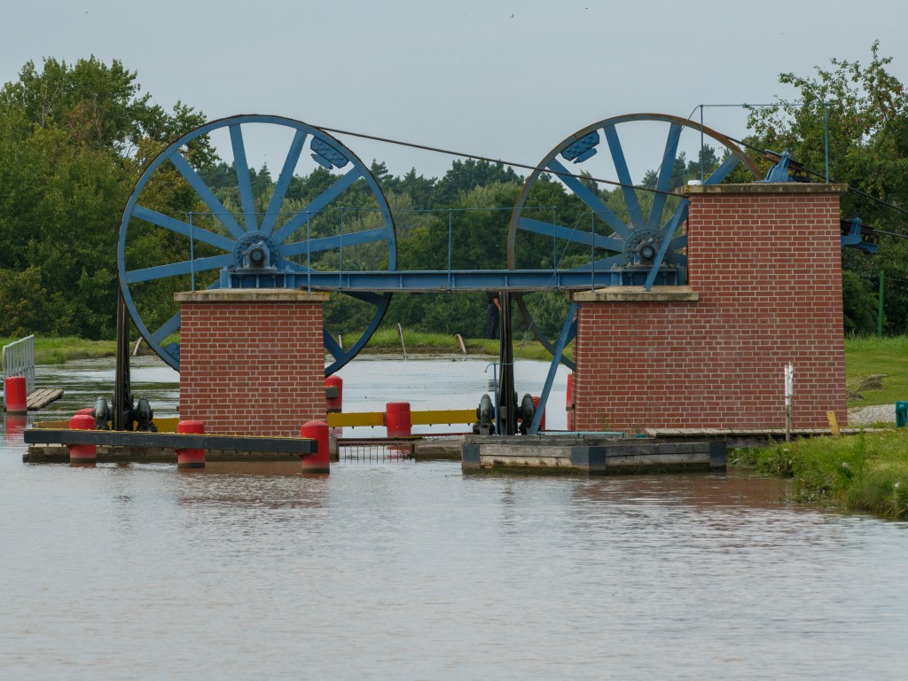 Mechanizm wodny z dużymi niebieskimi kołami zamontowany na murze ceglanym, z wodą w tle.